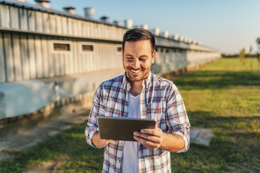 Cheerful caucasian farmer in plaid shirt standing outdoors and using tablet. In background are barns and orchard.
